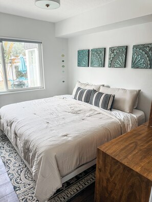 Ein Schlafzimmer mit Bett, einem Holfnachtkästchen, einem Fenster mit Blick und Wandbildern.