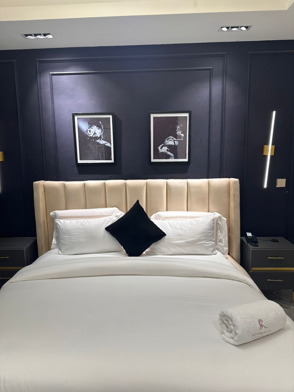 A neatly made bed with white linens and a black pillow, a beige headboard, and two framed pictures on the wall.