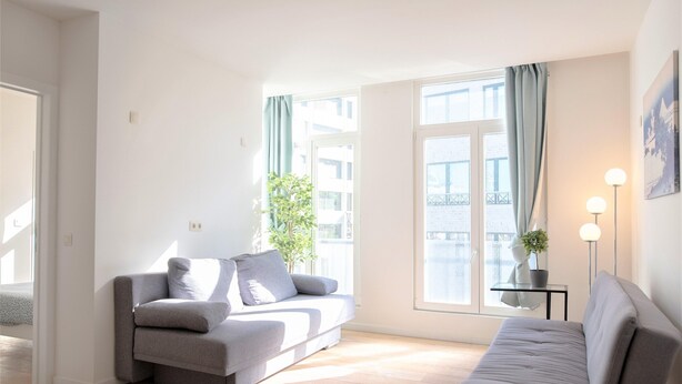 A modern living room with a grey sofa, a smaller grey armchair, a potted plant, and a floor lamp. - 12-guest Apartment on the Main Square (Antwerp)