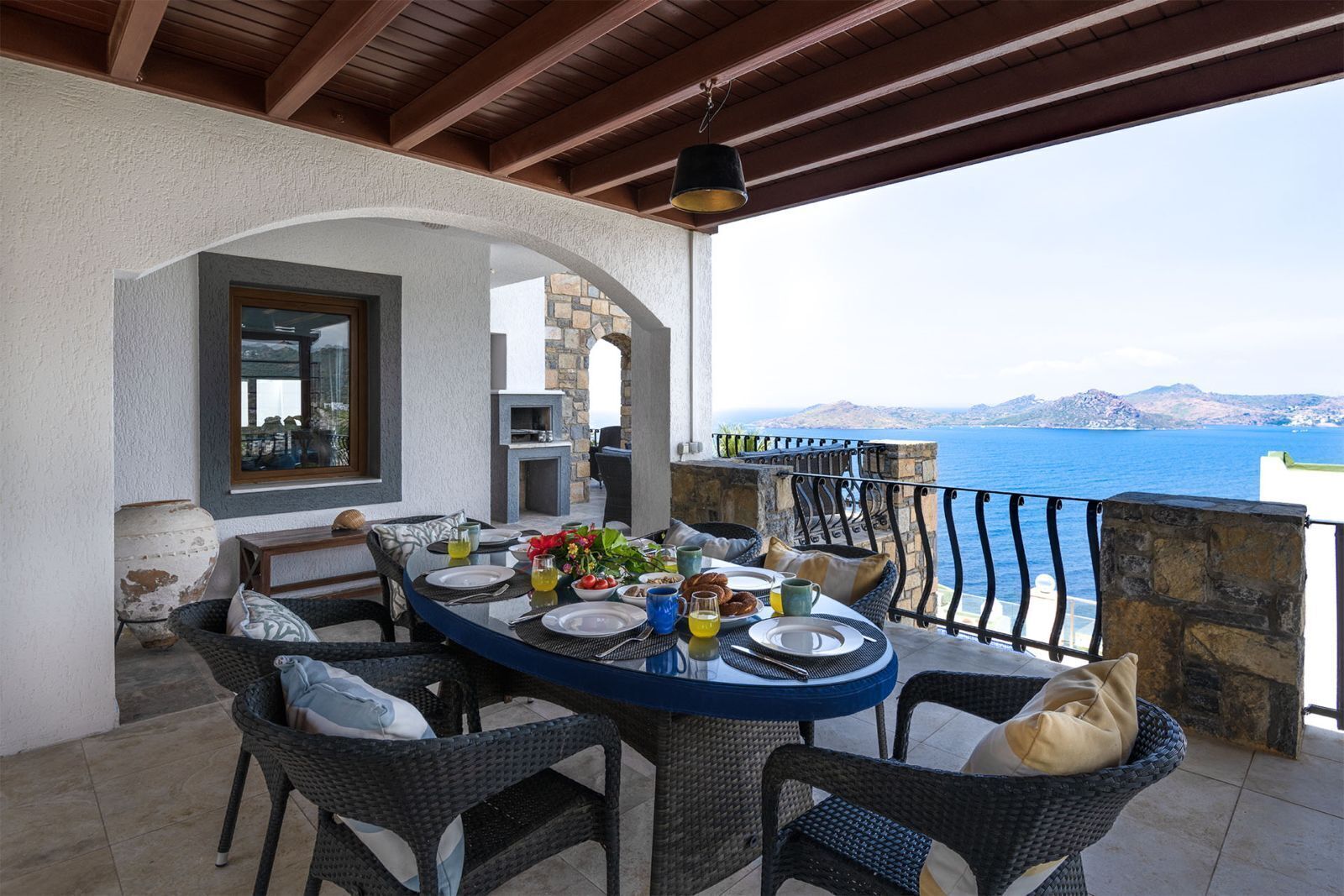 A covered outdoor dining area with a table set for two, a stone fireplace, and a view of the sea.