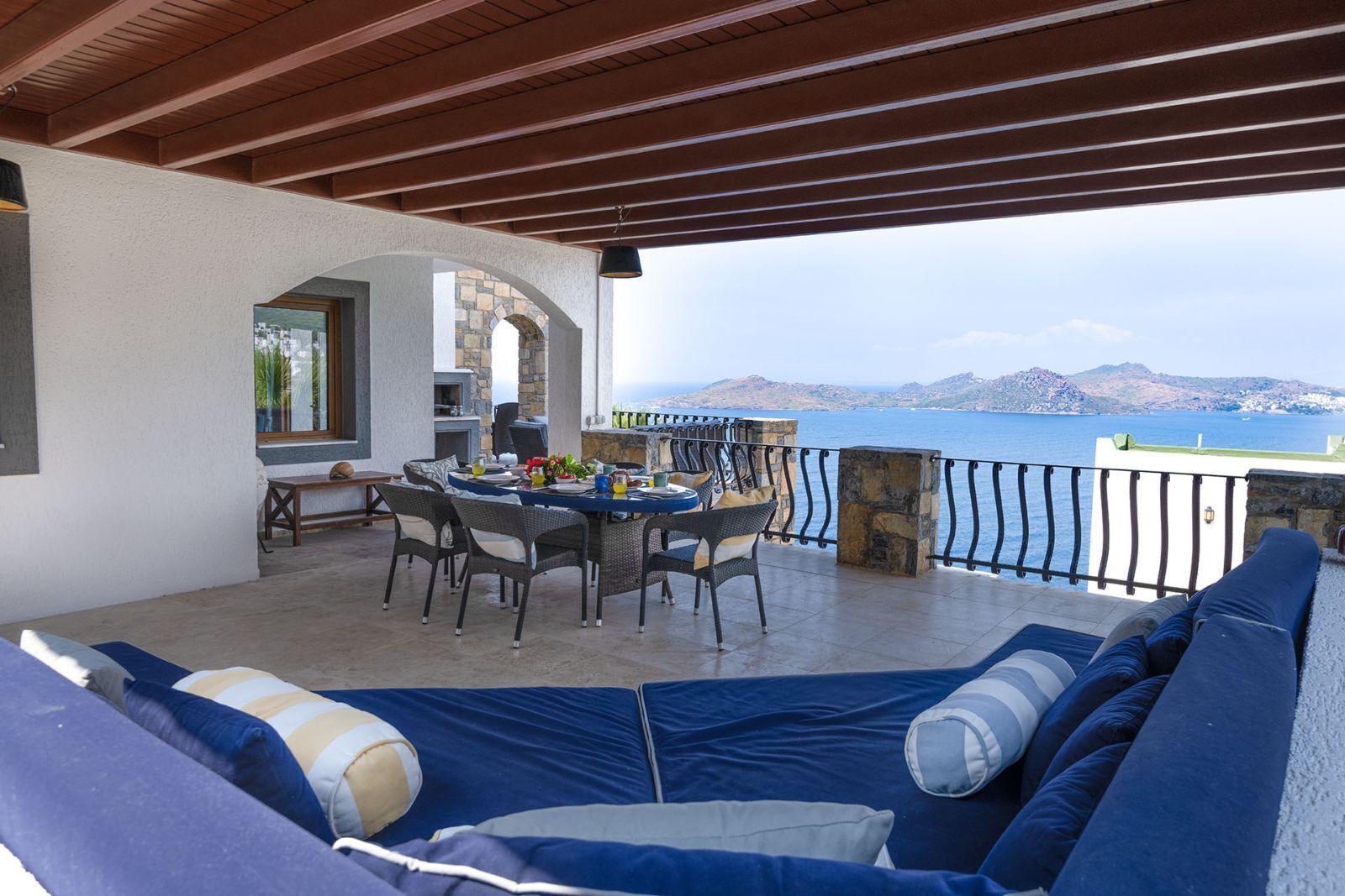A covered outdoor seating area with a table and chairs, a stone fireplace, and a view of the sea.