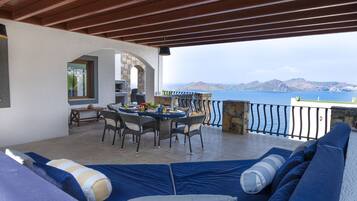 A covered outdoor seating area with a table and chairs, a stone fireplace, and a view of the sea.
