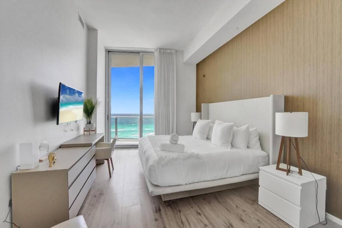 A modern hotel room with a bed, bedside table, lamp, and a TV on the wall.