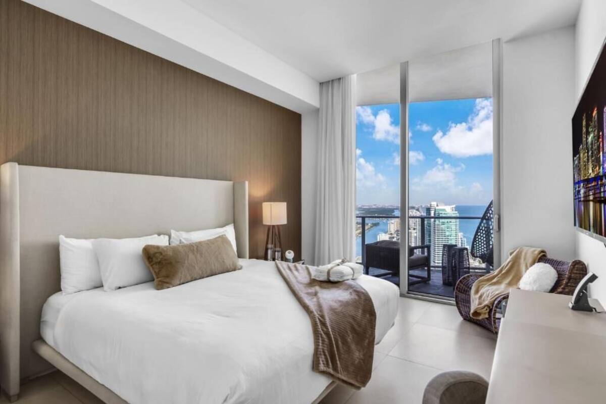 A hotel room with a large bed, a balcony with a city view, and a bookshelf.