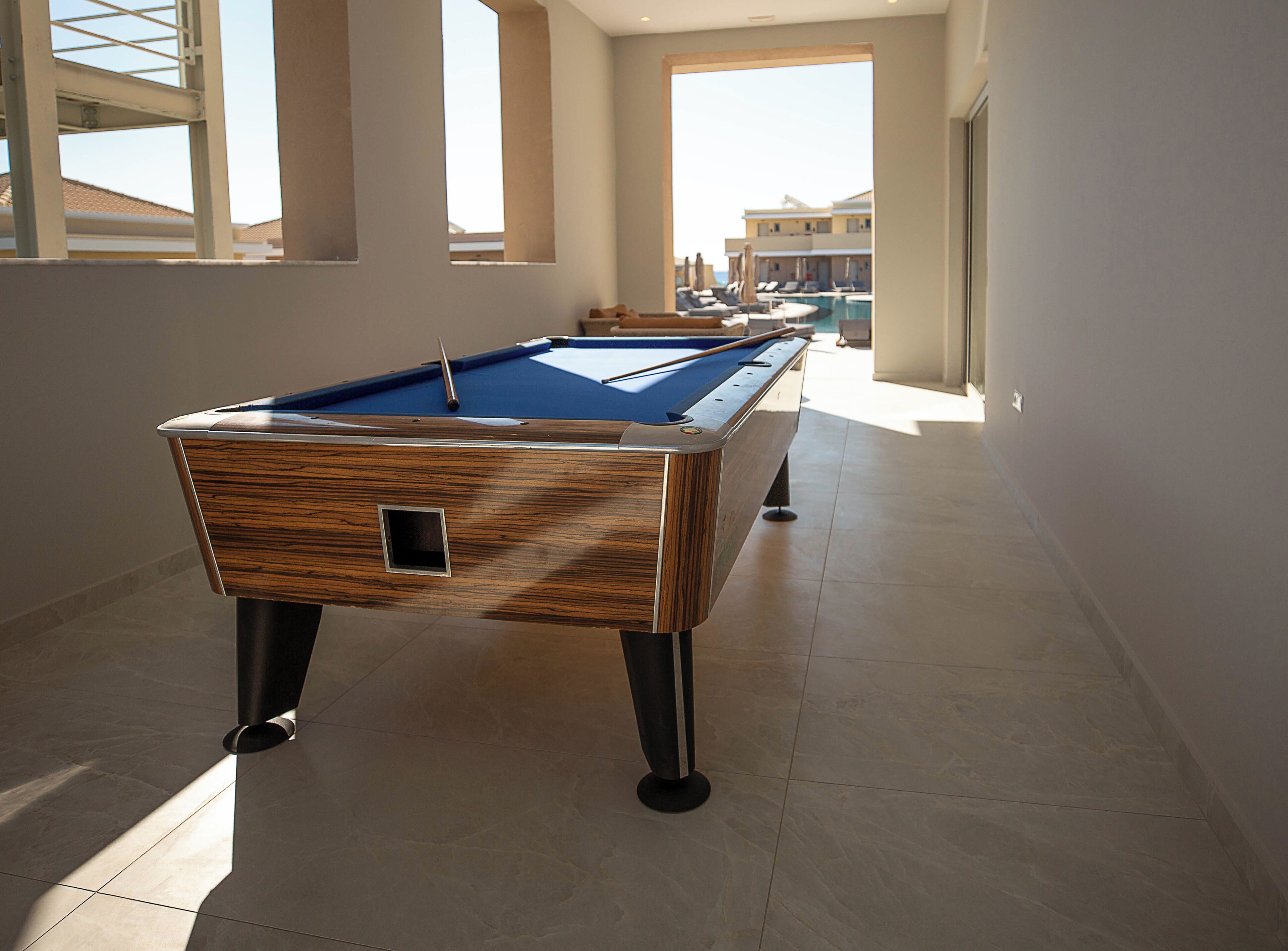 Una mesa de billar en un pasillo con vista al área de la piscina y edificios al fondo.