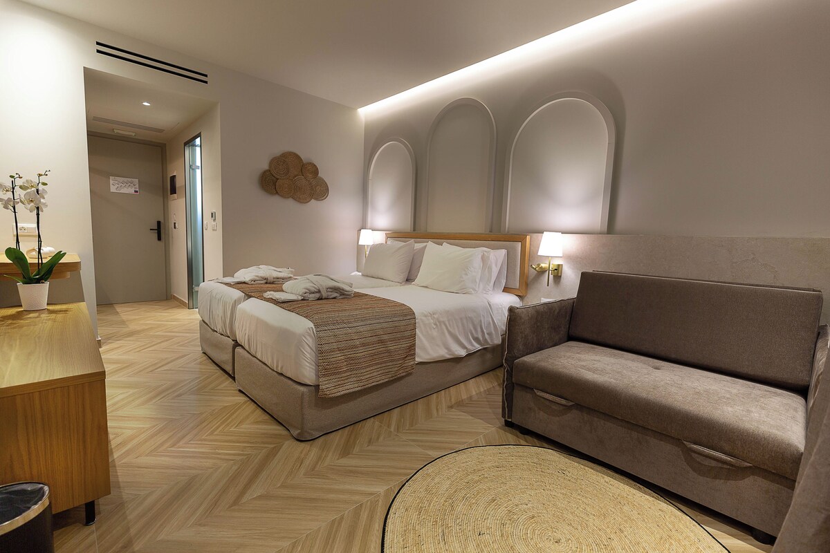 A modern hotel room with a bed, a sofa, a desk, and a potted plant.