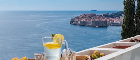 Ein Balkon mit Tablett, darauf ein Glas Wasser und eine Zitrone, mit Blick auf eine Küstenstadt mit historischen Gebäuden und blauem Meer.