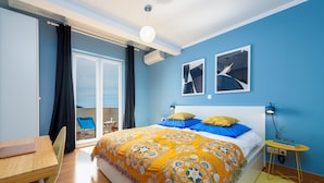 A bedroom with a bed, a desk, a chair, a small table with books and glasses, and a window with curtains.