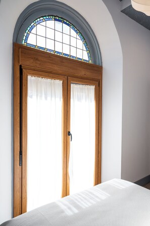 Una habitación con un amplio ventanal arqueado, una puerta de madera con un panel de vidrio satinado y una cama con una colcha blanca con textura.