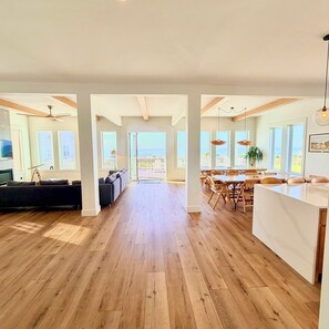 A spacious living area with a wooden floor, a dark sofa, and a dining area with a white island and wooden chairs.