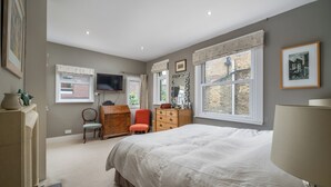 Ein Schlafzimmer mit einem großen Bett, einem Schminktisch, einem Sessel, einem Fernseher und einem Fenster mit Blick auf ein Backsteingebäude.
