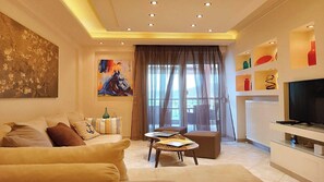 A modern living room with a sofa, coffee table, and a TV. There is a painting on the wall and a window with curtains. - Zeus Luxury Suite in Nea Flogita (Flogita)