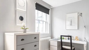 A room with a white desk, a chair, a radiator, a window with a black blind, and a framed picture on the wall. - Pavilion Road Mews House - 4 bed (London)
