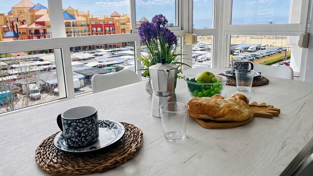 Taula amb una tassa, un got, un bol de fruita, un pastís i un ram de flors, amb vista a edificis i cotxes aparcats.