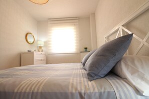 A neatly made bed with a striped bedspread, a bedside table with a lamp, a round mirror, and a window with blinds. - Carpe Diem by At Home Costa del Sol (Torrox)