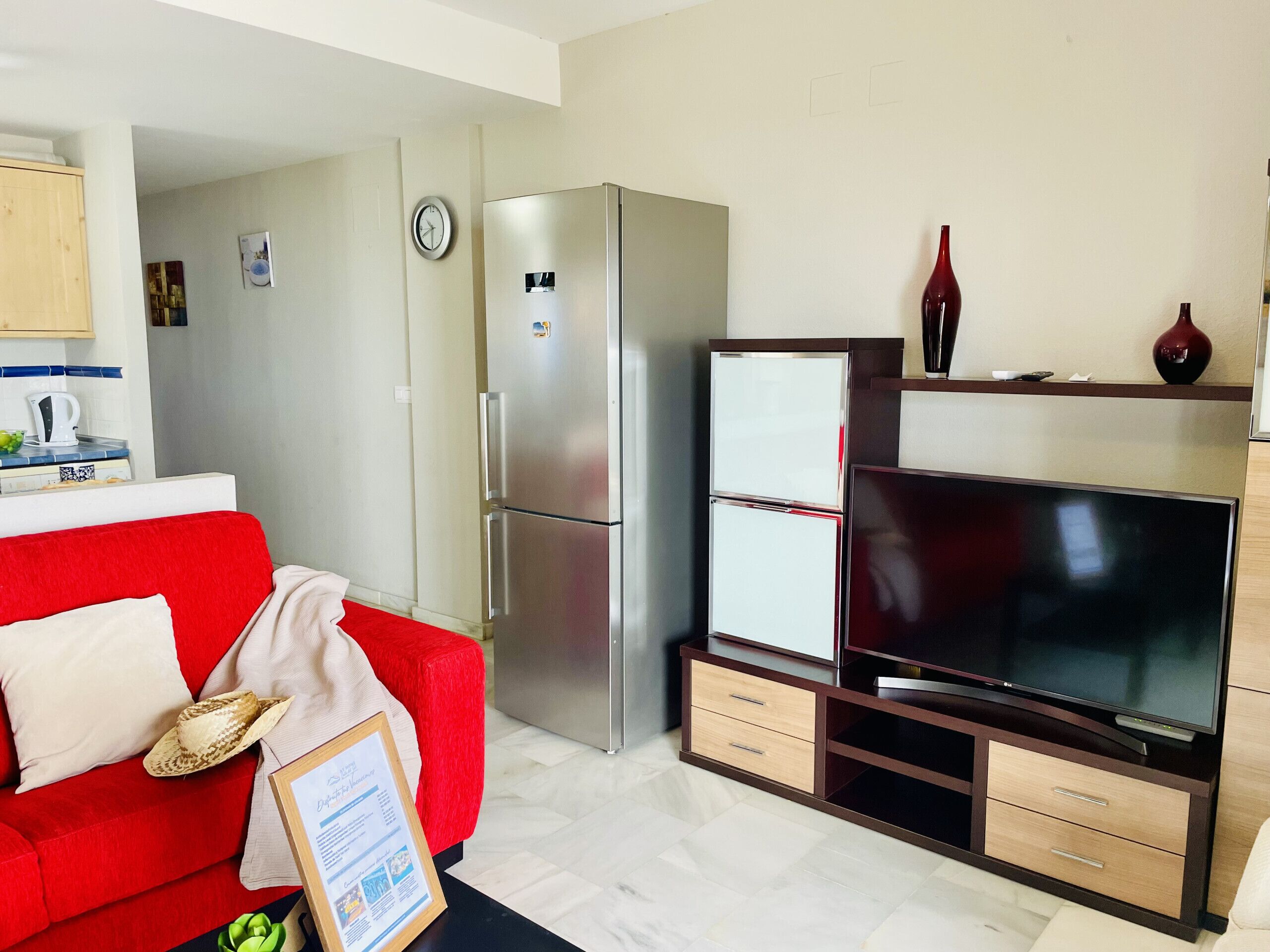 A living room with a red sofa, a flat-screen TV, a refrigerator, and a kitchen area with a kettle and a clock.