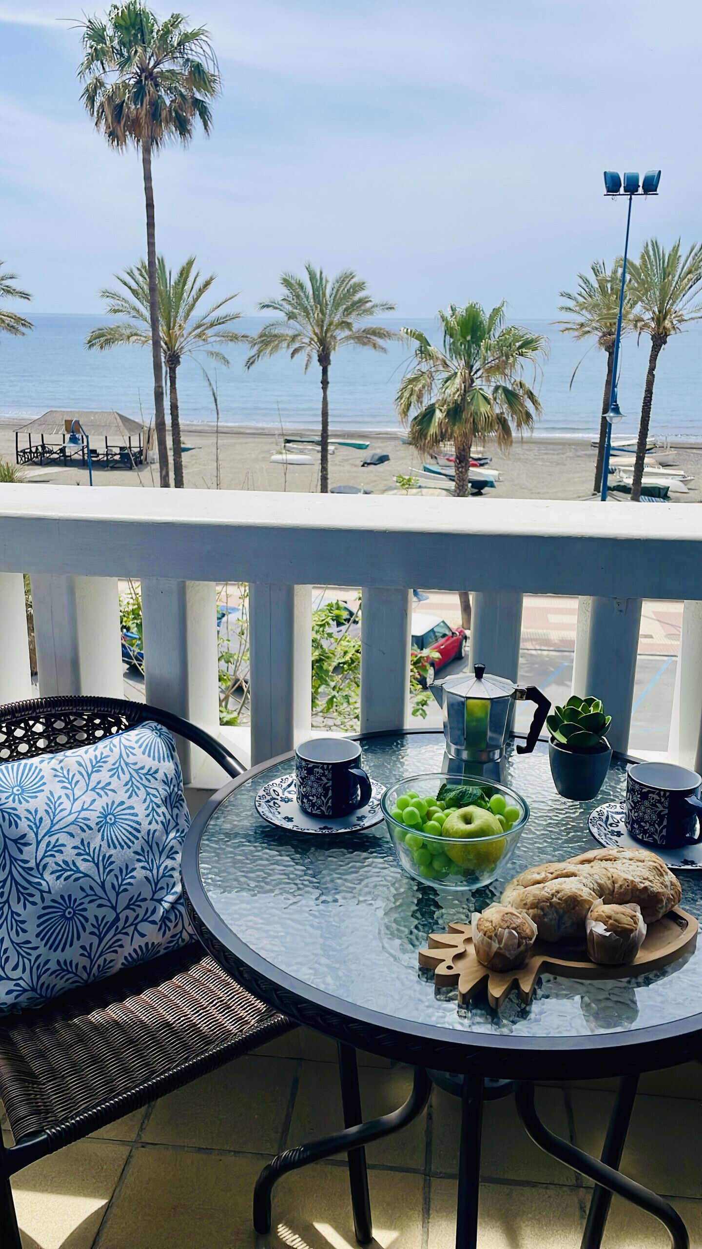 En balkon med et bord dækket til morgenmad, med udsigt over en strand med palmer og både.