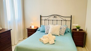 A neatly made bed with a green bedspread, a headboard, and multiple pillows. There are two bedside tables, one with a lamp and the other with a small plant. A wooden dresser is to the left. - Edificio Arrecife Primera L nea Vistas al Mar y (Torrox)