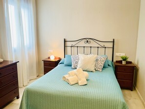 En pænt redt seng med grønt dynedække, en sengegavl og mange puder. To sengeborde, hvoraf det ene har en lampe og det andet en lille plante. En trækommode står til venstre.