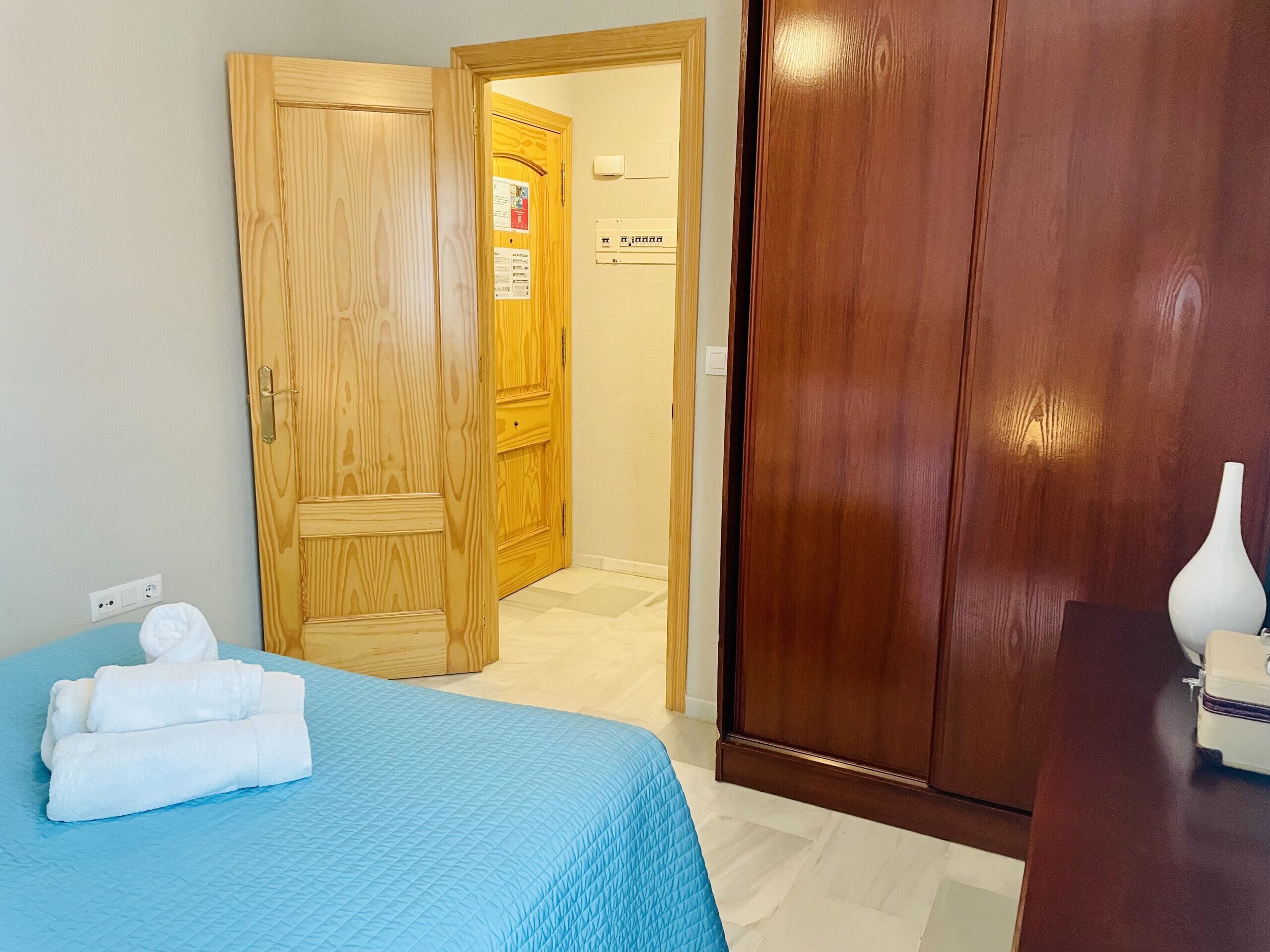 A hotel room with a bed, a wooden wardrobe, a door leading to another room, and a white vase on a table.