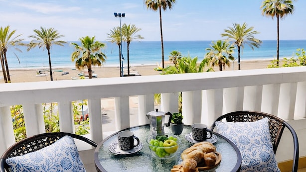 A balcony with a table set for two, overlooking a beach with palm trees and the ocean. - Edificio Arrecife Primera L nea Vistas al Mar y (Torrox)