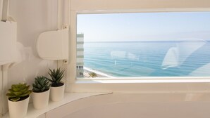 Une fenêtre donnant sur la mer, un mur blanc et trois plantes en pot.