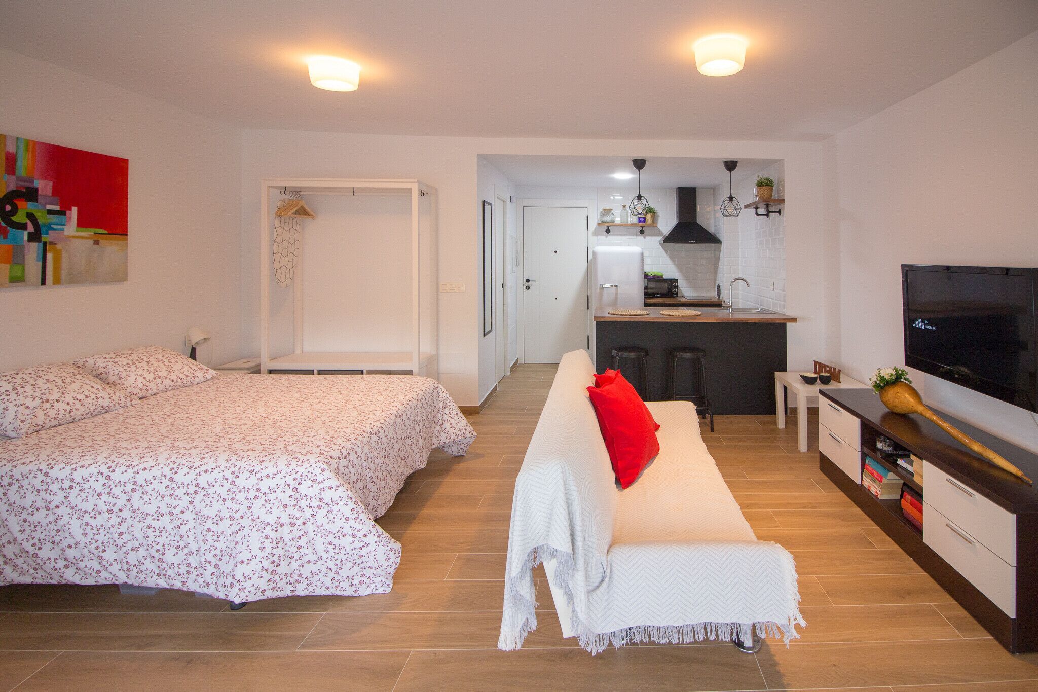 A modern apartment with a large bed, a sofa, a TV, and a kitchen area.