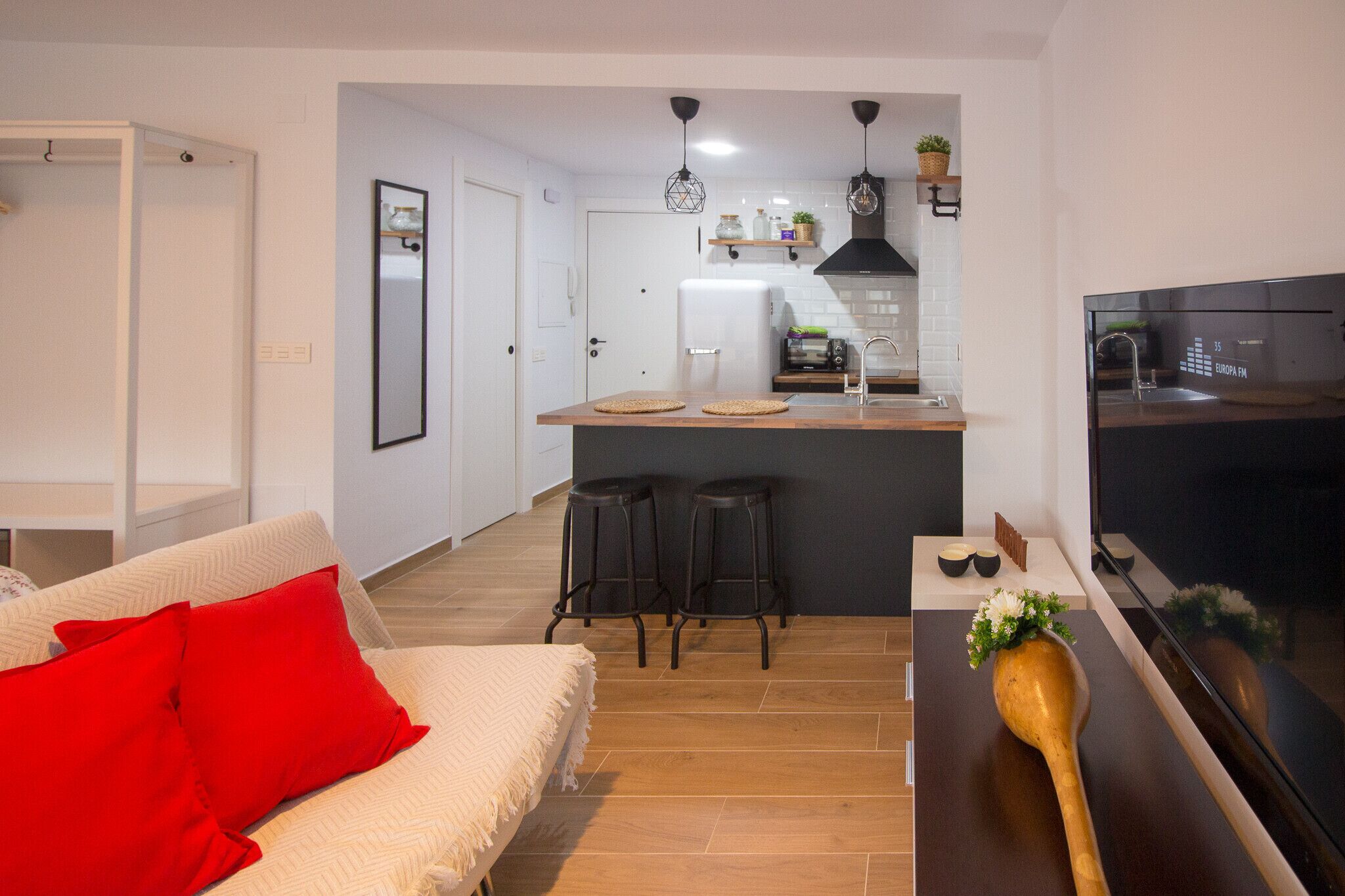 A modern kitchen with a central island, bar stools, a large flat-screen TV, and a dining area with a sofa and red cushions.