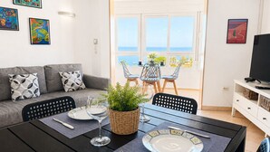 Een eethoek met een tafel gedekt voor twee personen, een bank met gestreepte kussens en uitzicht op de zee door grote ramen.
