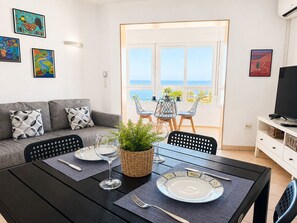 A dining area with a table set for two, a sofa with patterned cushions, and a view of the sea through large windows. - Madrid 401 by At Home Costa del Sol (Torrox)