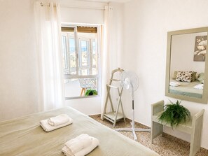 A hotel room with a bed, a standing fan, a chair, a small table, a mirror, and a window with curtains. - Madrid 401 by At Home Costa del Sol (Torrox)
