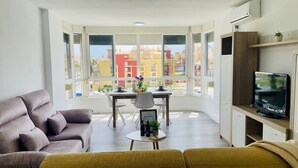 Ein modernes Wohnzimmer mit einer Couch, einem Esstisch mit Stühlen, einem Fernseher auf einem Holzgestell und großen Fenstern mit Blick auf farbenfrohe Gebäude.