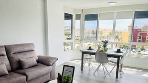 Ein modernes Wohnzimmer mit einem grauen Sofa, einem Esstisch mit Stühlen und einem großen Fenster mit Blick auf Gebäude und Palmen.