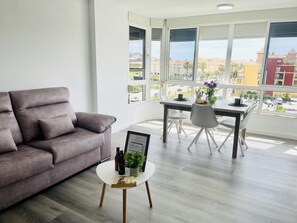 A modern living room with a grey sofa, a dining table with chairs, and a large window with a view of buildings and palm trees. - Laguna 967 by At Home Costa del Sol (Torrox)