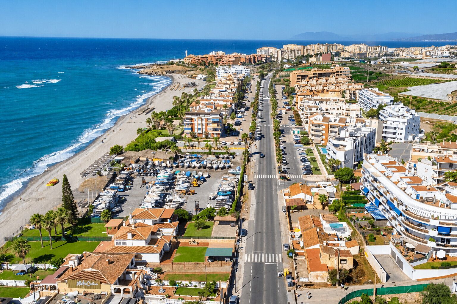 Vue aérienne d’une ville côtière avec une plage, une route principale et des immeubles résidentiels.