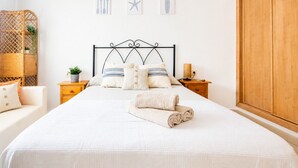 Een netjes opgemaakt bed met hoofdbord, een nachtkastje met lamp, een houten garderobe en een rieten mand met planten.