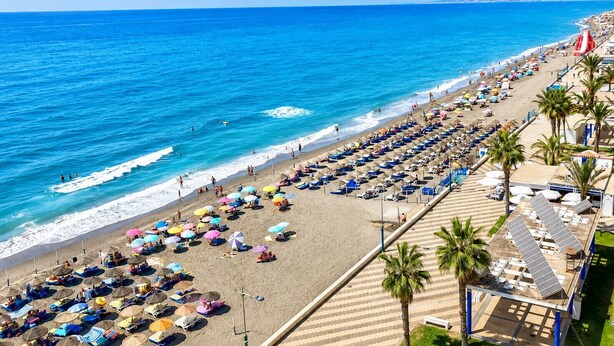Een strand met rijen ligbedden en parasols, een heldere blauwe zee en een verre kustlijn.