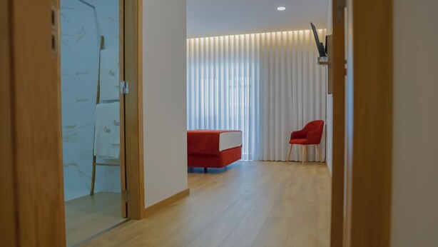 Una habitación de hotel moderna con piso de madera, un sofá rojo, una silla roja y un ventanal con persianas verticales.