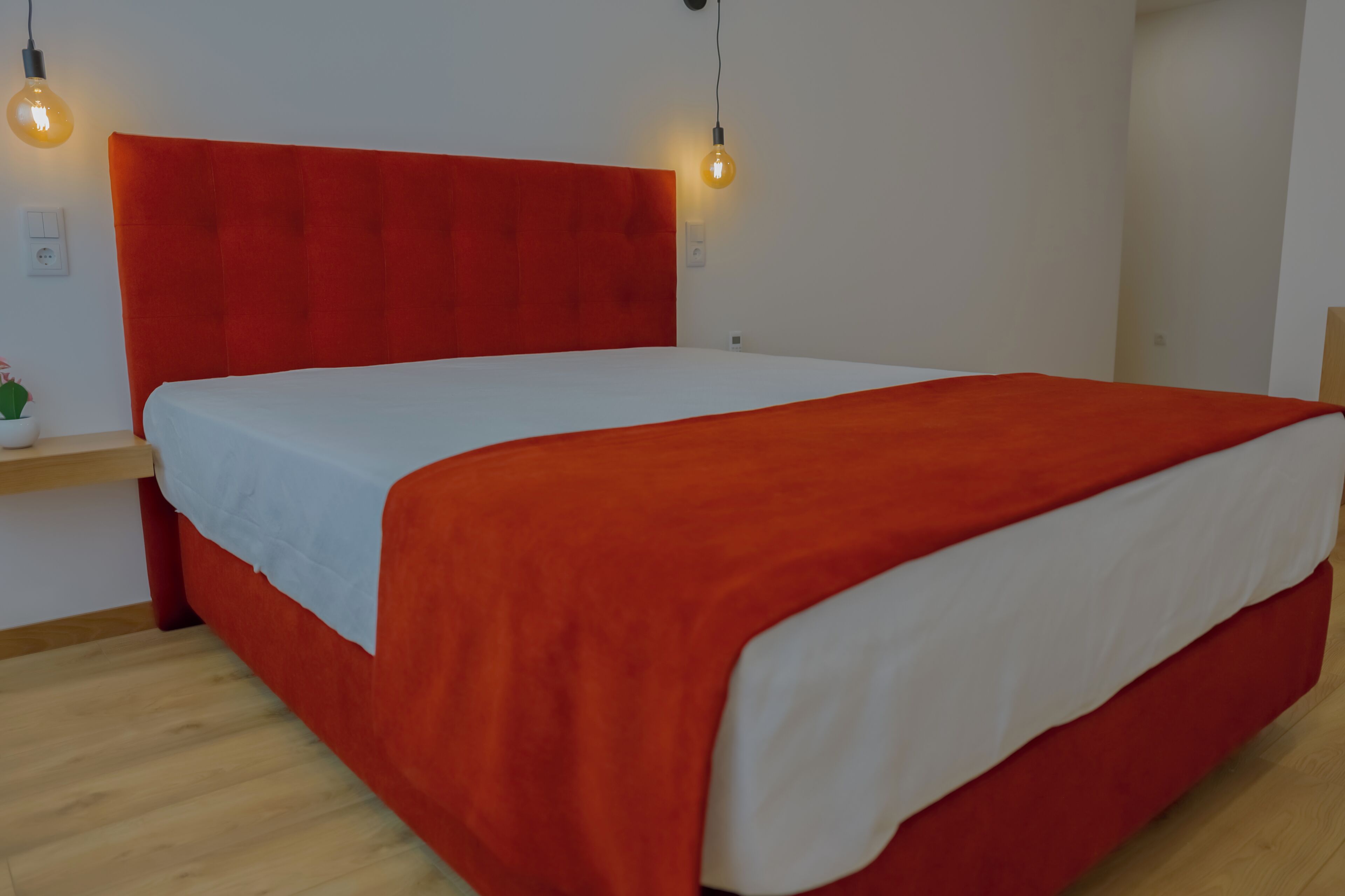 A bed with a red headboard and white bedding, a bedside plant, and two wall-mounted light bulbs.