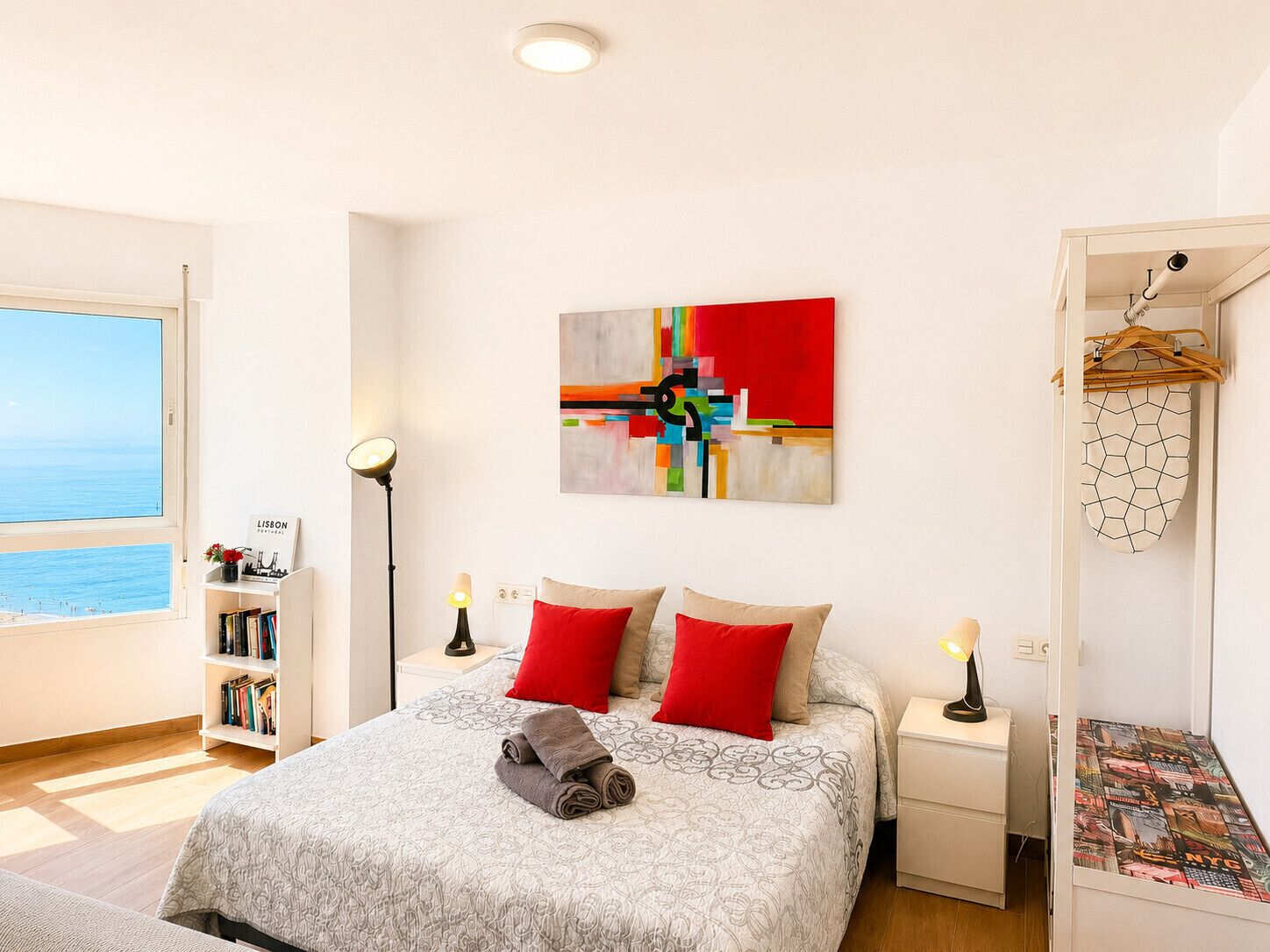 A bedroom with a bed, a nightstand, a bedside table, a bookshelf, and a view of the sea.