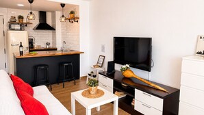 Een moderne keuken met een ingebouwde magnetron, een koffieapparaat en een spoelbak. Er is een eethoek met een witte tafel en een bank met rode kussens. Een televisie is aan de muur bevestigd en een houten lepel ligt op een kastje.