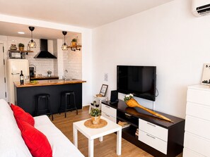 A modern kitchen with a built-in microwave, a coffee machine, and a sink. There is a dining area with a white table and a sofa with red cushions. A television is mounted on the wall, and a wooden spoon rests on a cabinet. - Madrid 810 by At Home Costa del Sol (Torrox)