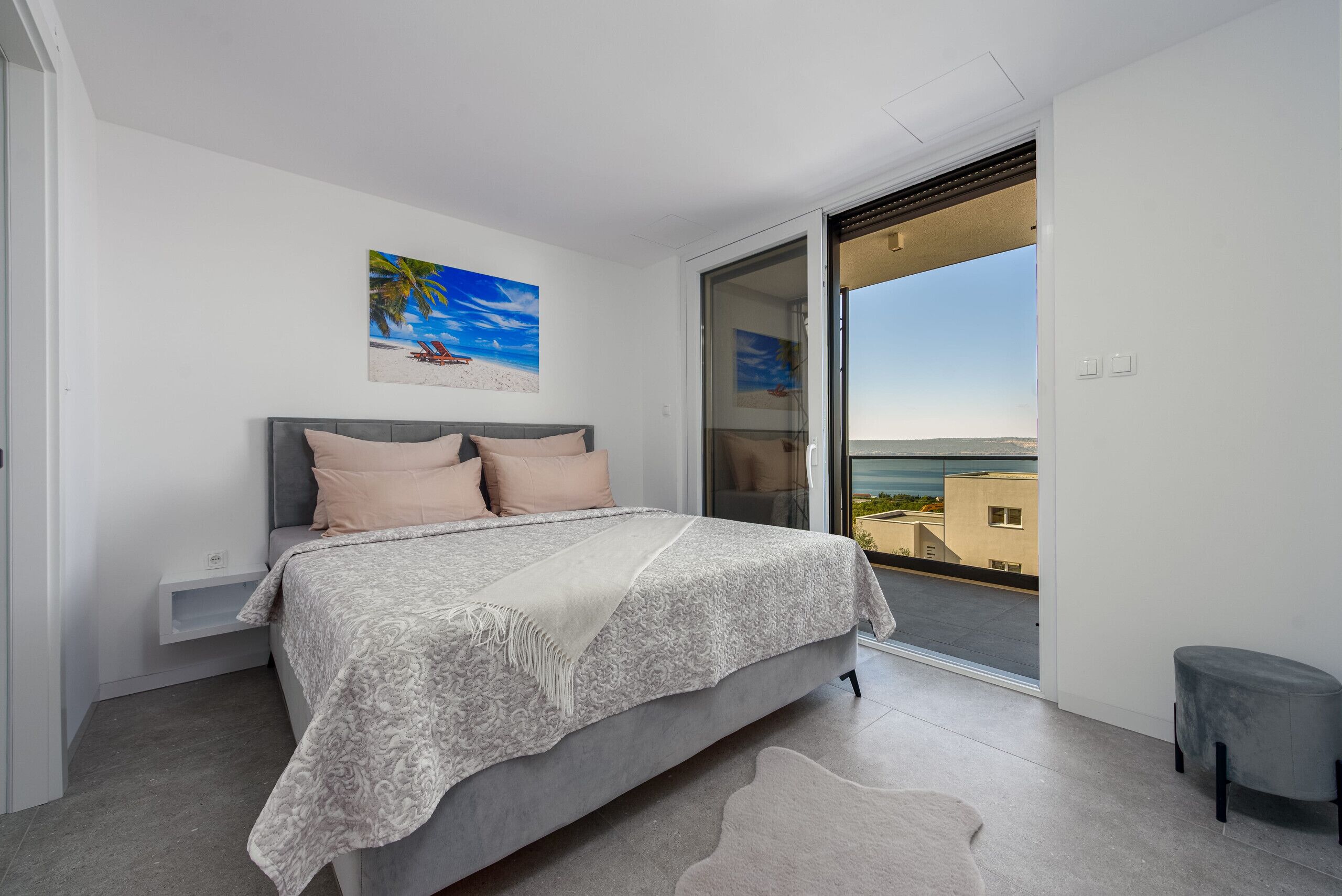 A bedroom with a bed, a nightstand, a painting, a sliding glass door to a balcony, and a small stool.