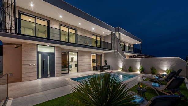 Una casa moderna de dos pisos con piscina, mobiliario exterior y un balcón bien iluminado.