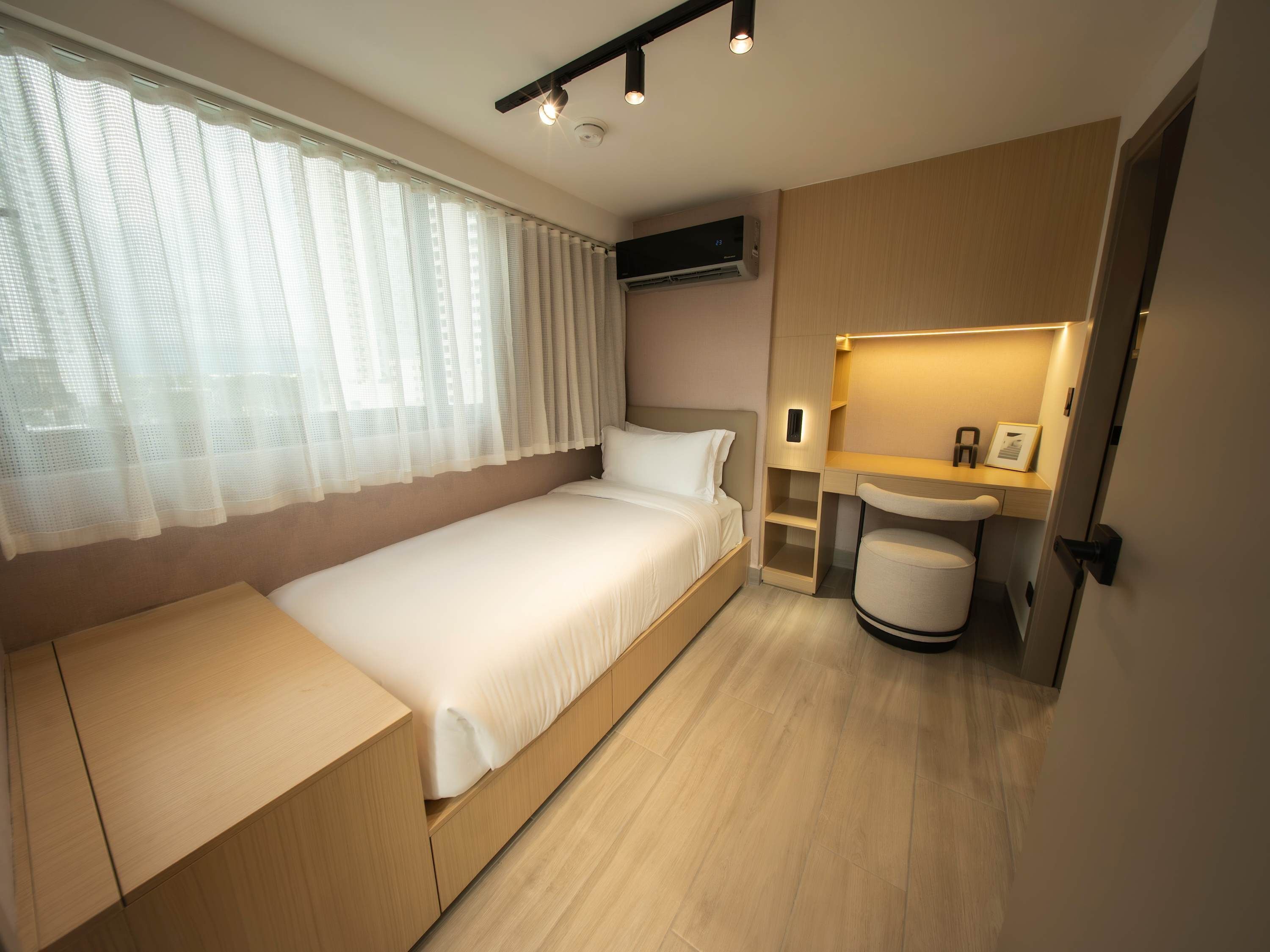 Habitación de hotel compacta con una cama individual, una mesita de noche pequeña, un banco integrado, una lámpara de noche, aire acondicionado instalado en la pared y un baño pequeño con lavabo y espejo.