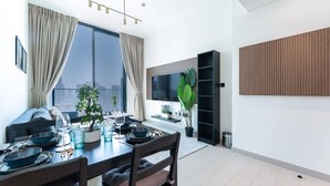 A modern dining area with a table set for four, a sofa, a TV, and a large window with curtains. - Free Housekeeping Visits During Your Stay - Stayshort - 1 Bed in Jumeirah Village Circle for 4 in a Great Location (Dubai)