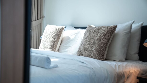 A neatly made bed with patterned pillows, a dark headboard, and a window with curtains. - Free Housekeeping Visits During Your Stay - Stayshort - 1 Bed in Jumeirah Village Circle for 4 in a Great Location (Dubai)