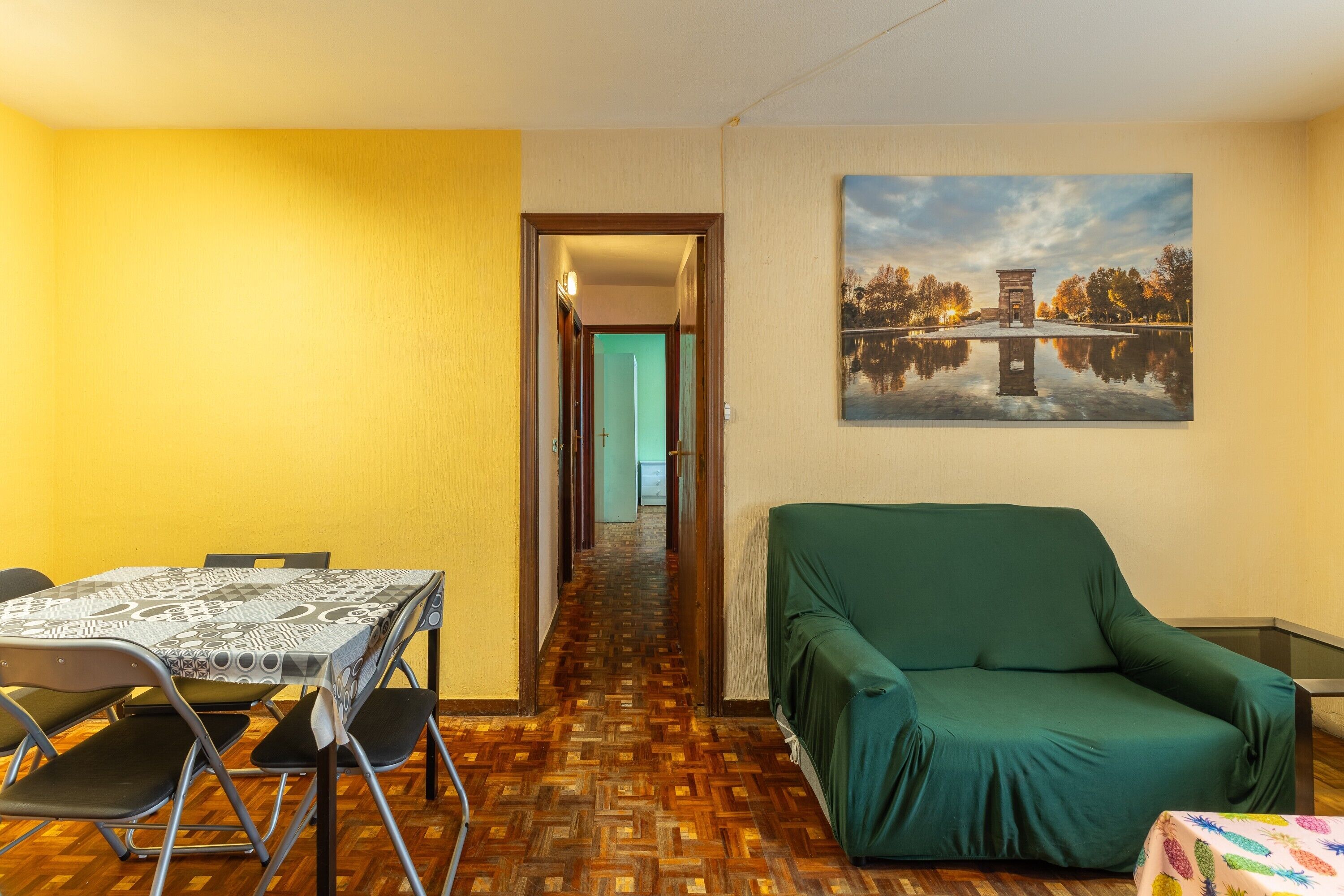A room with a wooden parquet floor, a green sofa, a dining table with chairs, and a painting on the wall.