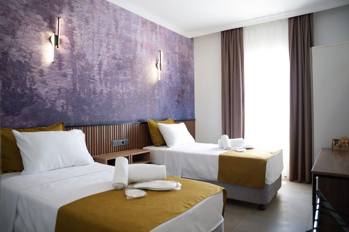 Habitación de hotel con dos camas, una mesita de noche de madera, un escritorio y un ventanal con cortinas.