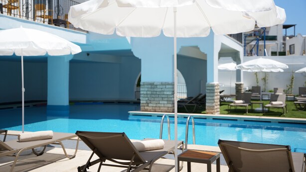 Un área junto a la piscina con sillones de descanso, sombrillas blancas y una piscina de color azul cristalino.